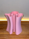 A light pink sculptural pencil holder with a wavy, fluted silhouette, standing on a wooden surface against a neutral wall with a warm light strip.