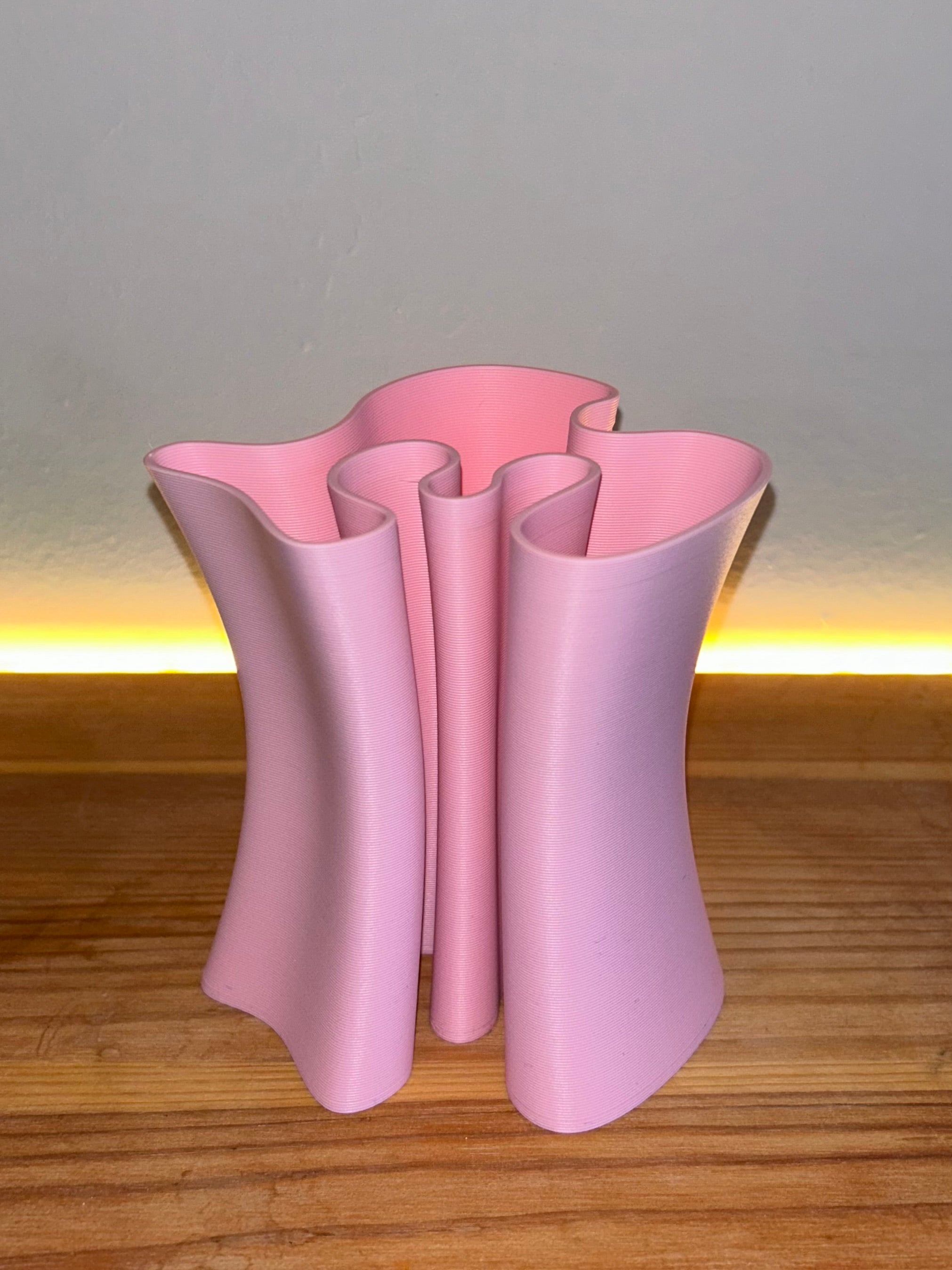 A light pink sculptural pencil holder with a wavy, fluted silhouette, standing on a wooden surface against a neutral wall with a warm light strip.